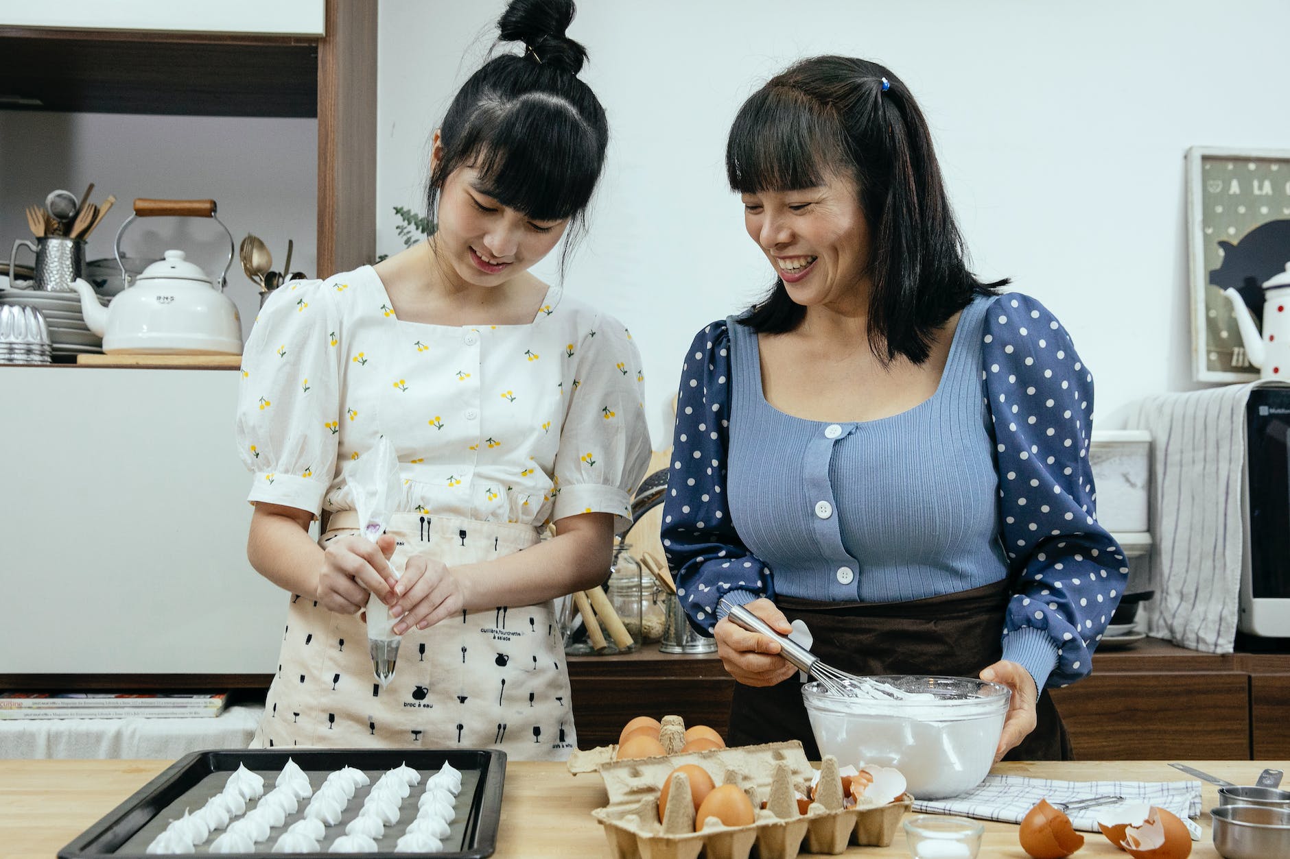 cheerful young ethnic lady with mother cooking meringue together in kitchen